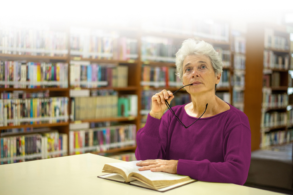 Titelbild der Literaturtage LeseZeichen 2025 - es zeigt eine Frau, sitzend am Tisch in der Bibliothek. Sie hat ein Buch aufgeschlagen und lässt ihren Blick noch oben schweifen.
