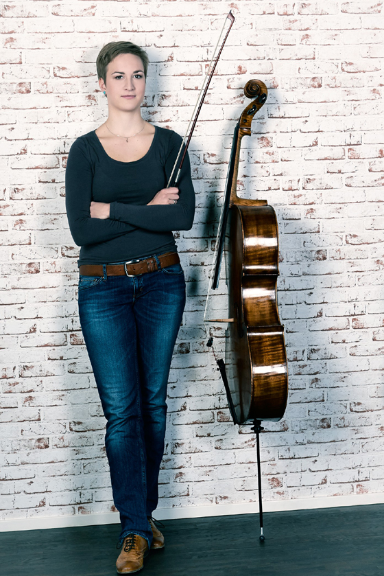Das Bild zeigt Ulrike Münch stehend vor einer Ziegelwand. Sie hält ihren Bogen in der Hand und neben ihr steht ihr Violoncello. Ulrike Münch ist Musiklehrerin für Violoncello an der Musikschule Mittelsachsen.