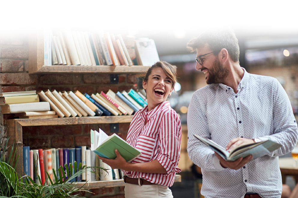 Titelbild der Literaturtage LeseZeichen 2026 - es zeigt eine Frau und einen Mann. Beide stehen vor einem Bücherregal, halten ein geöffnetes Buch in der Hand und schauen sich lachend an.