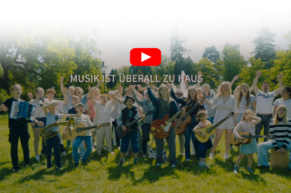 Ausschnitt des Image-Videos der Musikschule Mittelsachsen, bei dem viele Kinder und Erwachsene musizieren und singen. Alle stehen auf einer grünen Wiese bei Sonnenschein.